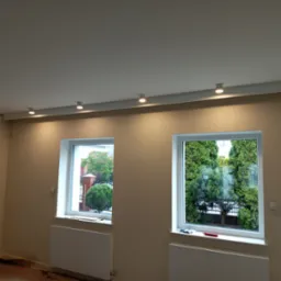 Jasne wnętrze z dwoma oknami i rzędem okrągłych lamp sufitowych LED, świecących ciepłym światłem, zamontowanych na listwie przy suficie. Ściany w odcieniach beżu.