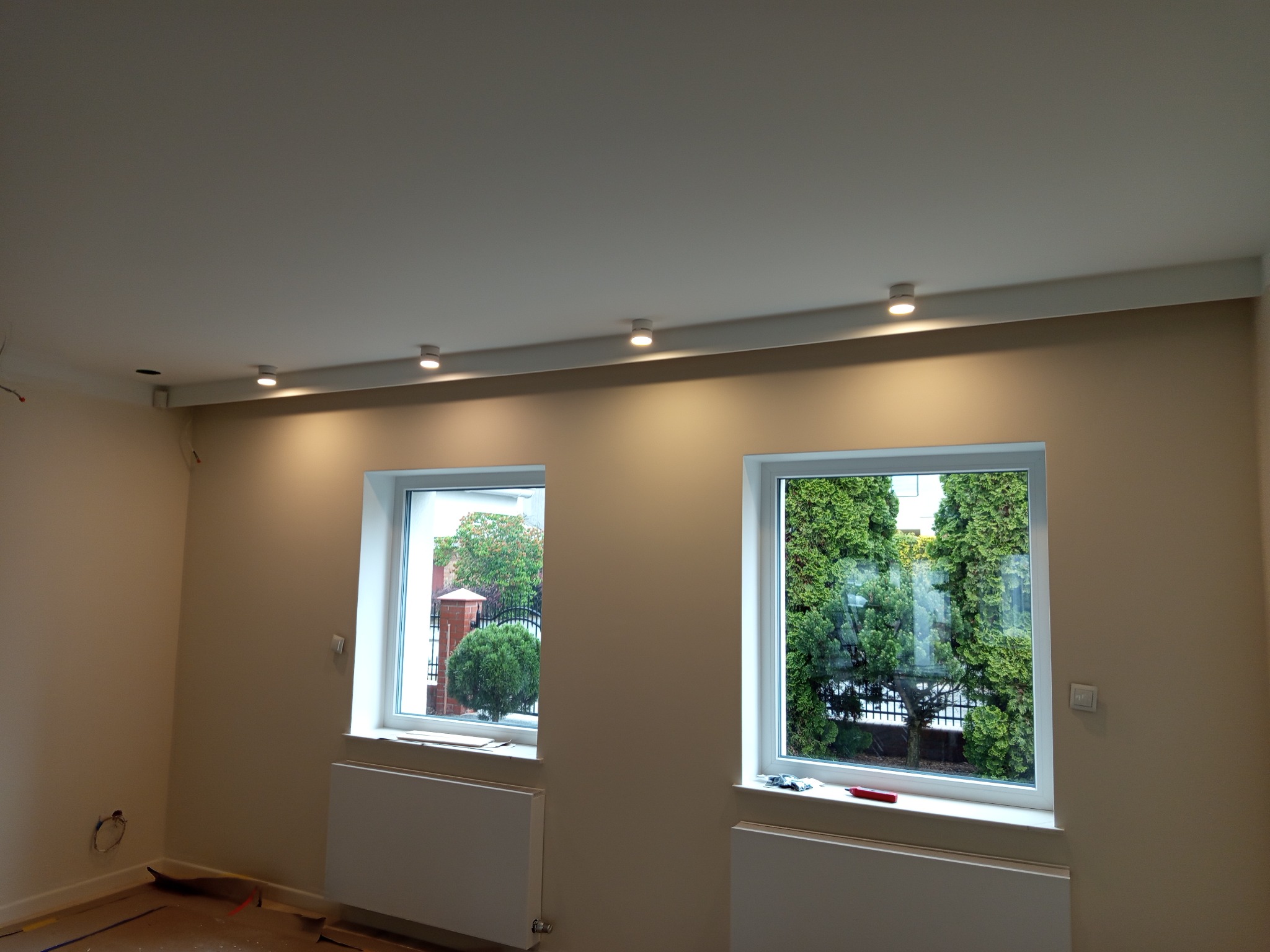 Jasne wnętrze z dwoma oknami i rzędem okrągłych lamp sufitowych LED, świecących ciepłym światłem, zamontowanych na listwie przy suficie. Ściany w odcieniach beżu.