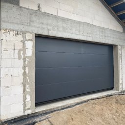 Finestre Rafał Łuczak - Zamknięta, segmentowa brama garażowa w kolorze grafitowym, zamontowana w surowym stanie budynku z białego bloczka. Widoczne detale montażu i otoczenie budowy.