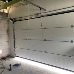 Finestre Rafał Łuczak - Jasna brama garażowa segmentowa, widok od wewnątrz, z widocznymi elementami montażowymi i narzędziami. Ściany surowe, w trakcie wykańczania.