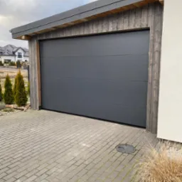 Nowoczesna, segmentowa brama garażowa w kolorze grafitowym, z drewnianym obramowaniem, na tle posesji z kostką brukową i budynkiem w tle.