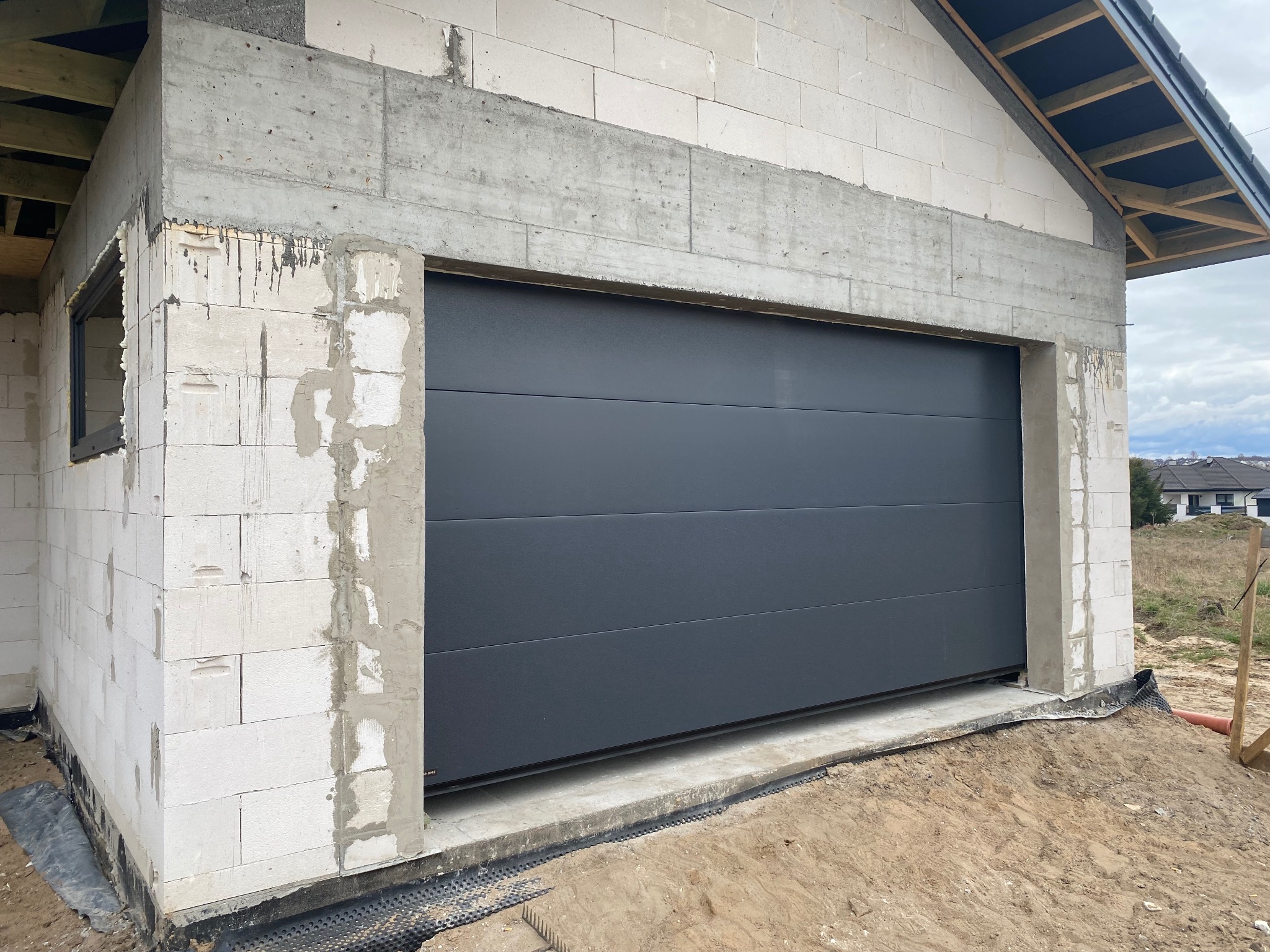 Zamknięta, segmentowa brama garażowa w kolorze grafitowym, zamontowana w surowym stanie budynku z białego bloczka. Widoczne detale montażu i otoczenie budowy.