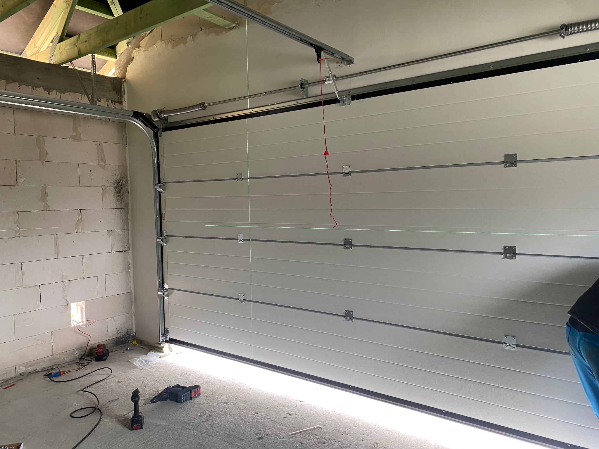 Jasna brama garażowa segmentowa, widok od wewnątrz, z widocznymi elementami montażowymi i narzędziami. Ściany surowe, w trakcie wykańczania.