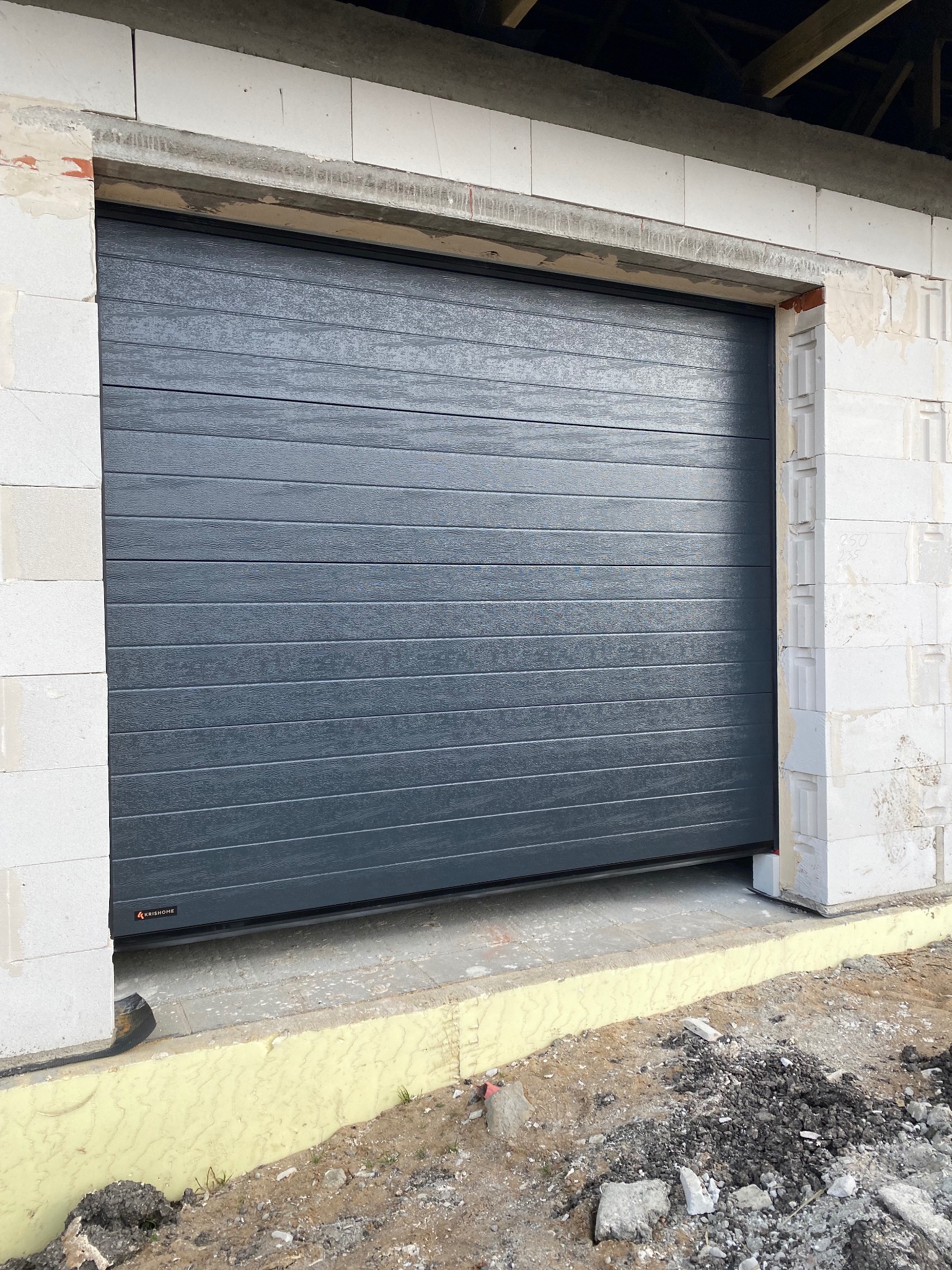 Szara, segmentowa brama garażowa KRISHOME, zamontowana w surowym otworze budynku z betonu komórkowego. Widoczne detale montażowe i fragment fundamentu.