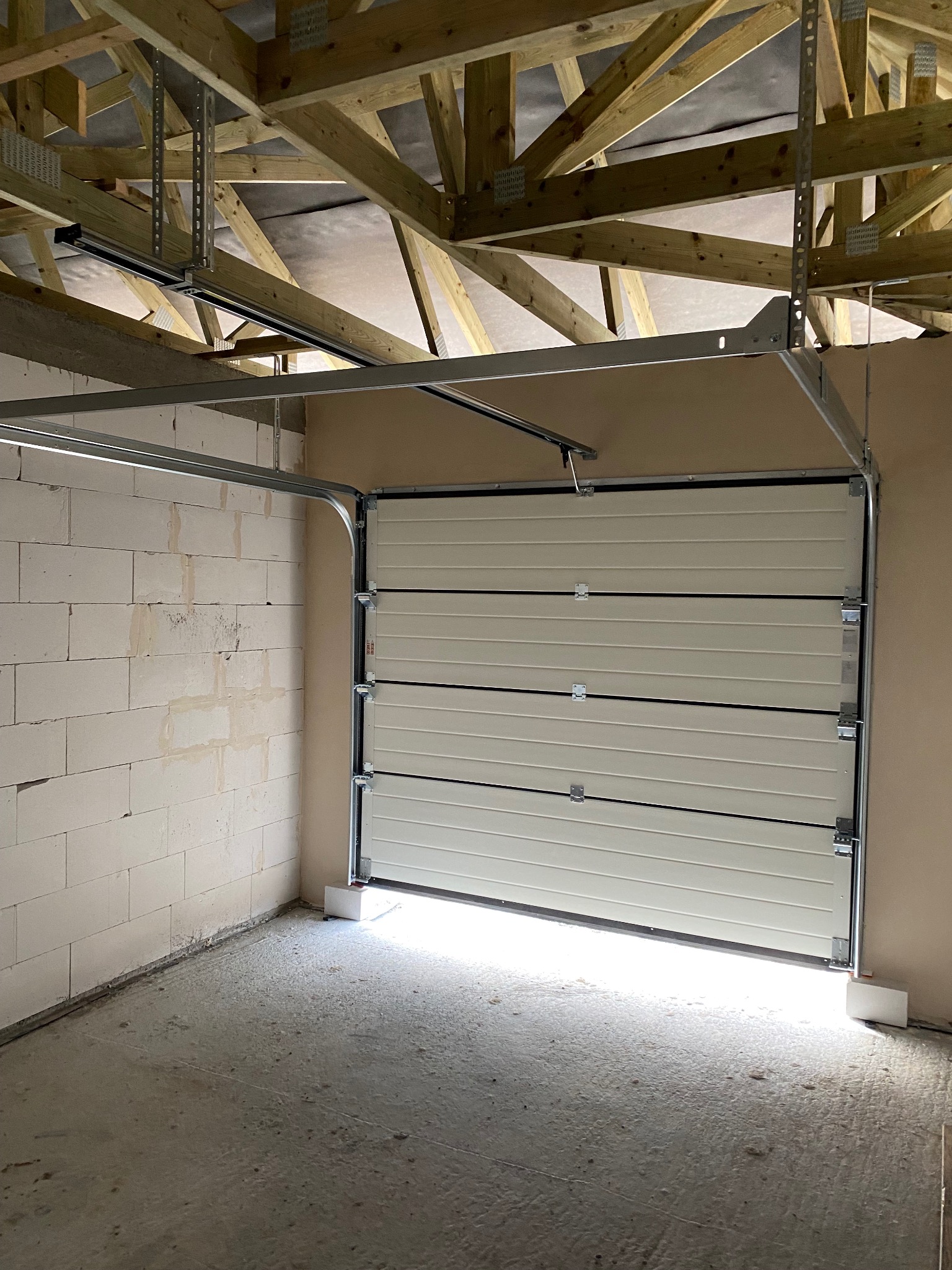 Otwarta segmentowa brama garażowa w surowym wnętrzu garażu z widoczną konstrukcją drewnianą dachu i ścianami z betonu komórkowego.