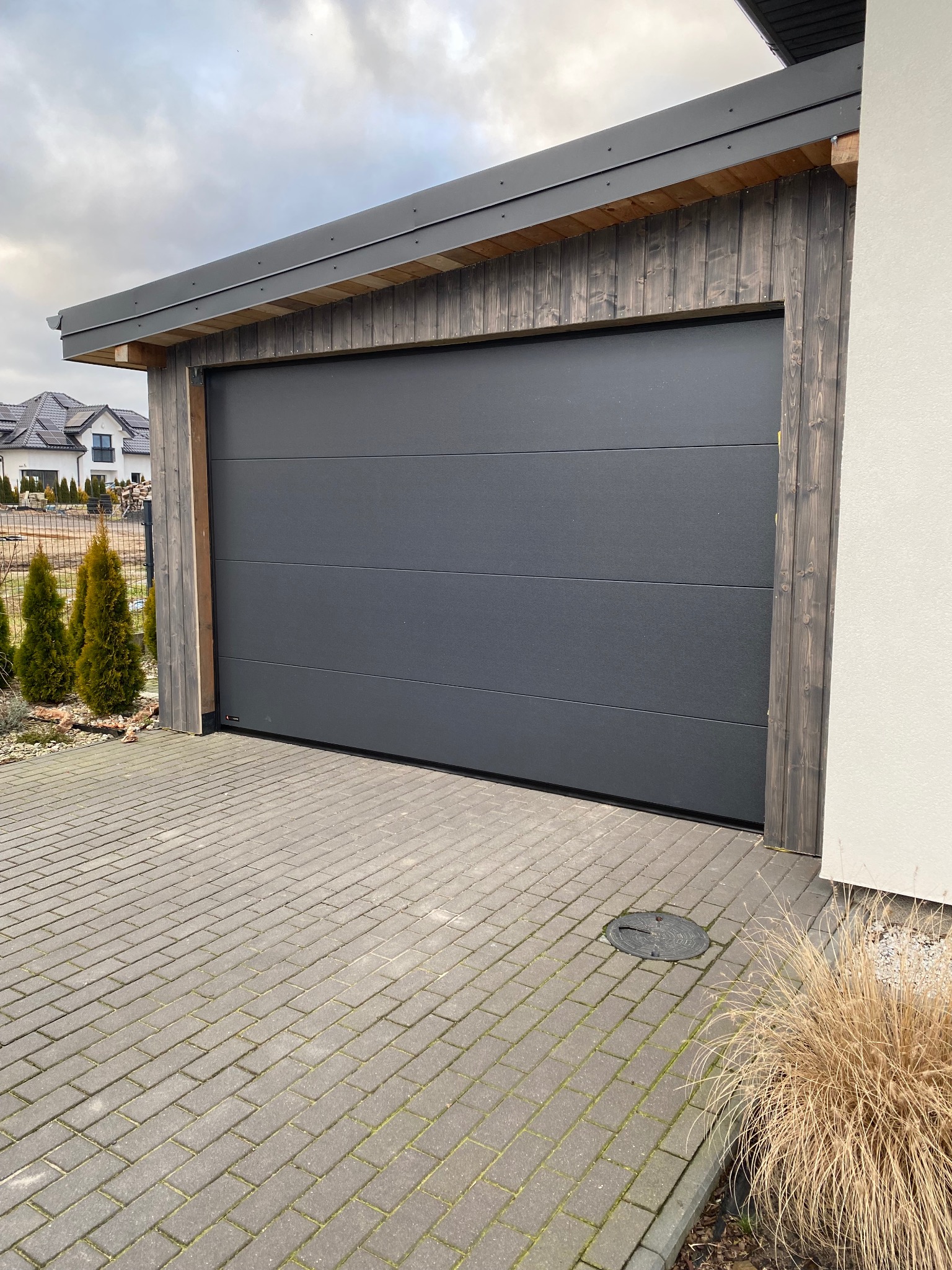 Nowoczesna, segmentowa brama garażowa w kolorze grafitowym, z drewnianym obramowaniem, na tle posesji z kostką brukową i budynkiem w tle.