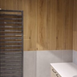 REFORMA REMONT W FORMIE SPÓŁKA Z OGRANICZONĄ ODPOWIEDZIALNOŚCIĄ - Wykończona łazienka z drewnopodobnymi płytkami na ścianie, szarym grzejnikiem drabinkowym i białą umywalką z szafką. Nowoczesny design.