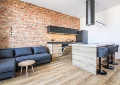 Loftowa kuchnia z ceglaną ścianą, drewnianą podłogą i wyspą. Czarny okap cylindryczny. Szare krzesła barowe i sofa. Nowoczesny design z elementami industrialnymi.