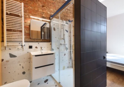 Nowoczesna łazienka z kabiną prysznicową, umywalką z szafką i grzejnikiem na tle ceglanej ściany. Uwagę przykuwają płytki imitujące lastryko i czarna ściana z płytek.