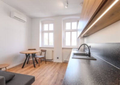 Jasne wnętrze z aneksem kuchennym: czarny blat, drewniane szafki z oświetleniem LED, okrągły stół i krzesła. Minimalistyczny design z klimatyzacją.