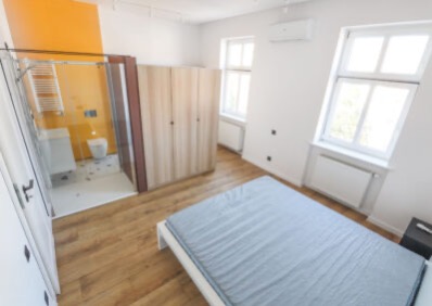 Jasne wnętrze z łazienką oddzieloną szklaną ścianą, widoczna szafa i łóżko. Żółta ściana w łazience dodaje koloru. Drewniana podłoga i białe ściany tworzą nowoczesny wygląd.