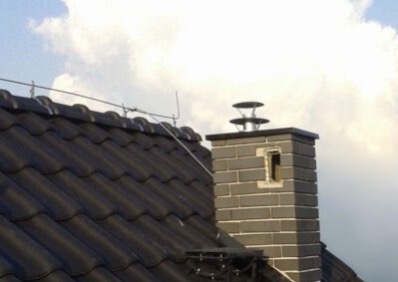 Grafitowa dachówka ceramiczna z systemem odgromowym i kominem z cegły klinkierowej na tle pochmurnego nieba. Widok z dołu.