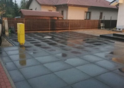 Szklana podłoga tarasu z widoczną konstrukcją nośną, żółta skrzynka elektryczna, ogrodzenie z brązowych desek w tle. Nietypowe rozwiązanie architektoniczne.