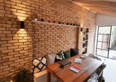 Ceglana ściana z oświetleniem i półkami na rośliny, drewniany stół z laptopem i poduszkami w tle, Poznań. Wykończenie wnętrza z charakterem.