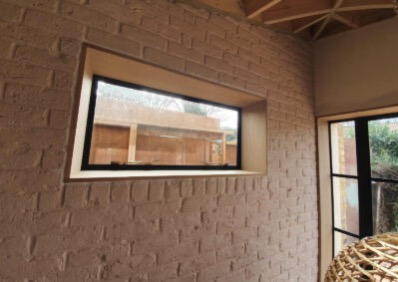Ściana z cegły pomalowanej na beżowo z oknem w czarnej ramie. Widoczny fragment sufitu z drewnianych belek i fragment drugiego okna. Nietypowa faktura ściany.