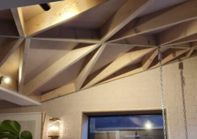 Nowoczesny sufit z drewnianą konstrukcją kratownicową i oświetleniem punktowym, widok z dołu, ściana z cegły pomalowana na biało, okno w tle.