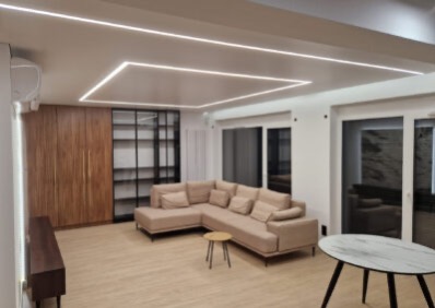 Jasne wnętrze salonu z narożnikiem, drewnianą zabudową i oświetleniem LED w suficie. Widoczne okna balkonowe i stoliki kawowe. Minimalistyczny design.