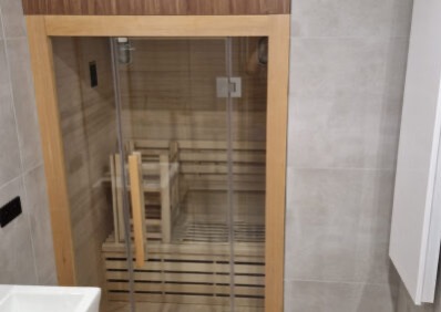 Nowoczesna sauna w łazience z drewnianym wykończeniem i szklanymi drzwiami, widoczna na tle szarych płytek. Minimalistyczny design.
