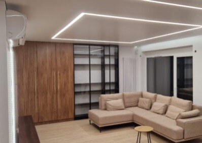 Nowoczesny salon z narożnikiem, drewnianą zabudową i podświetlanym sufitem. Minimalistyczny design, jasne kolory, widok na okno. Oświetlenie LED podkreśla geometrię wnętrza.