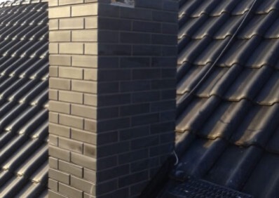 Ciemny, murowany komin z cegły na tle dachu pokrytego ciemną dachówką. Widok z dołu, podkreślający solidność konstrukcji i precyzję wykonania.