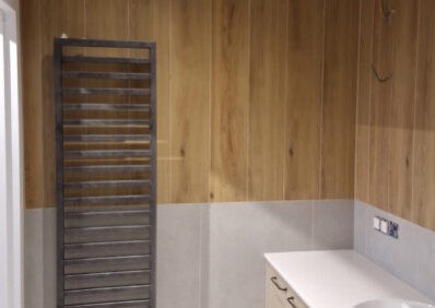 Wykończona łazienka z drewnopodobnymi płytkami na ścianie, szarym grzejnikiem drabinkowym i białą umywalką z szafką. Nowoczesny design.