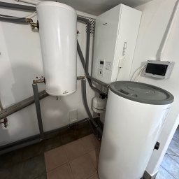 HELIUS ENERGY SPÓŁKA Z OGRANICZONĄ ODPOWIEDZIALNOŚCIĄ - Biała pompa ciepła Galmet i zasobnik Rotenso w kotłowni. Szare rury, płytki podłogowe w odcieniach brązu. Widoczny panel sterowania Panasonic.
