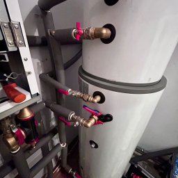 HELIUS ENERGY SPÓŁKA Z OGRANICZONĄ ODPOWIEDZIALNOŚCIĄ - Biały, dwuczęściowy zasobnik ciepłej wody użytkowej z rurami w izolacji szarej, z zaworami z czerwonymi motylkami, zamontowany w pomieszczeniu technicznym.