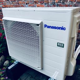 HELIUS ENERGY SPÓŁKA Z OGRANICZONĄ ODPOWIEDZIALNOŚCIĄ - Biała jednostka zewnętrzna klimatyzacji Panasonic R32 na tle ceglanej ściany i zieleni. Widoczne detale montażu na czarnych podkładkach i metalowych wspornikach.