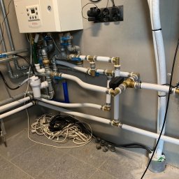 HELIUS ENERGY SPÓŁKA Z OGRANICZONĄ ODPOWIEDZIALNOŚCIĄ - Instalacja grzewcza z kotłem gazowym, rurami z zaworami i przewodami elektrycznymi na tle szarej ściany. Widoczne elementy instalacji w pomieszczeniu technicznym.