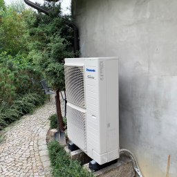 HELIUS ENERGY SPÓŁKA Z OGRANICZONĄ ODPOWIEDZIALNOŚCIĄ - Zewnętrzna jednostka klimatyzacji Panasonic Inverter, zamontowana na betonowym podeście przy budynku z szarą elewacją. Obok ścieżka z kostki i zieleń.