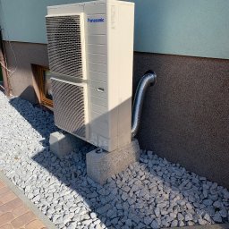HELIUS ENERGY SPÓŁKA Z OGRANICZONĄ ODPOWIEDZIALNOŚCIĄ - Zewnętrzna jednostka klimatyzacji Panasonic zamontowana na betonowych podstawach, z widocznym przewodem odprowadzającym skropliny. Otoczenie wyłożone ozdobnym kamieniem.