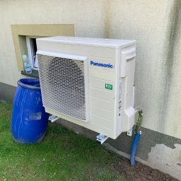 HELIUS ENERGY SPÓŁKA Z OGRANICZONĄ ODPOWIEDZIALNOŚCIĄ - Zamontowany klimatyzator Panasonic na elewacji budynku w Częstochowie. Widoczna niebieska beczka obok urządzenia i fragment okna.