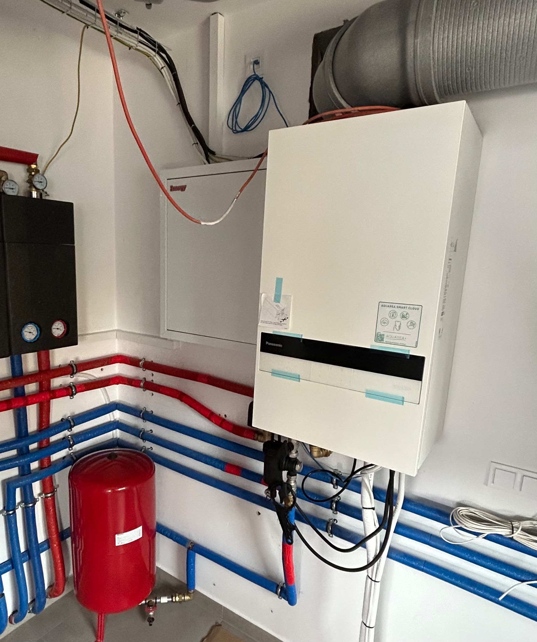Nowoczesna instalacja pompy ciepła Panasonic Aquarea z czerwonym zbiornikiem wyrównawczym i rurami w kolorach czerwonym i niebieskim, zamontowana w schludnym pomieszczeniu.