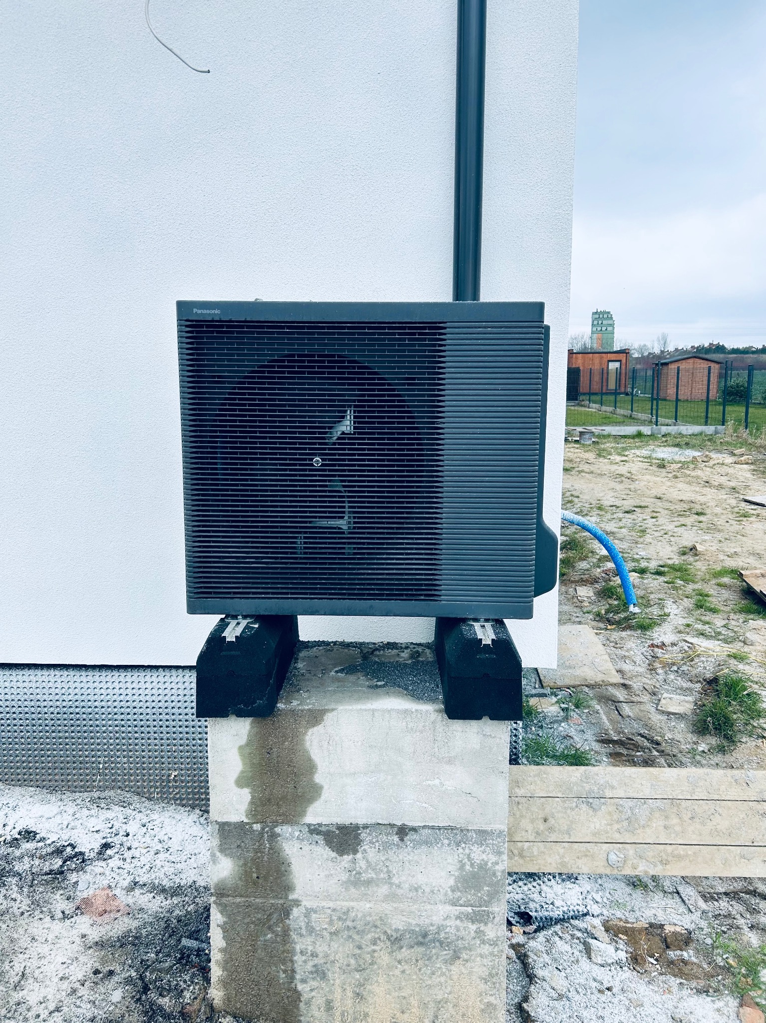 Zewnętrzna jednostka pompy ciepła Panasonic, zamontowana na betonowym fundamencie przy białej ścianie budynku. Widoczny fragment instalacji i teren budowy w tle.