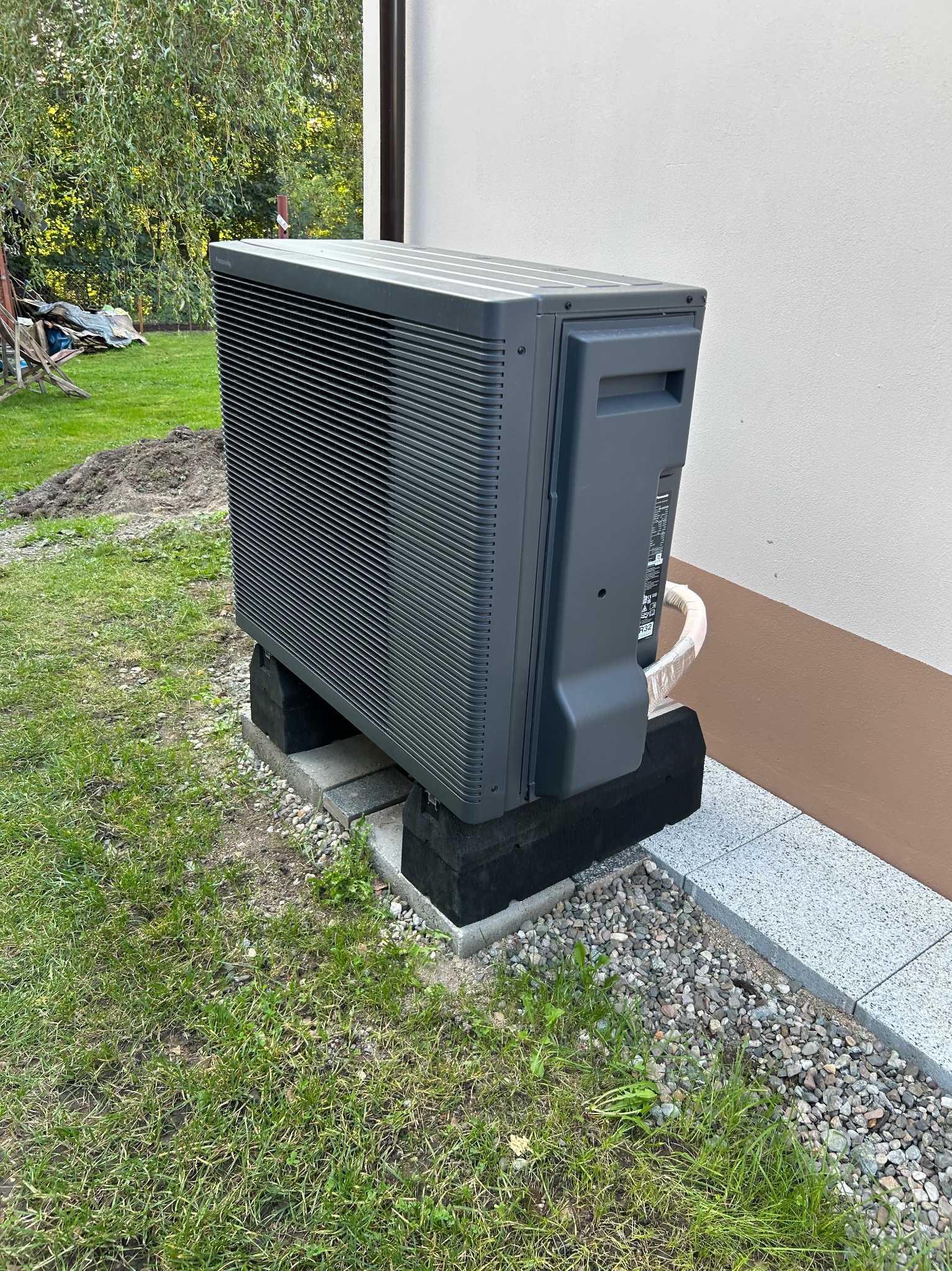 Szara pompa ciepła Panasonic Aquarea zainstalowana na zewnątrz budynku, na betonowych bloczkach, z widocznym fragmentem trawnika i elewacji.