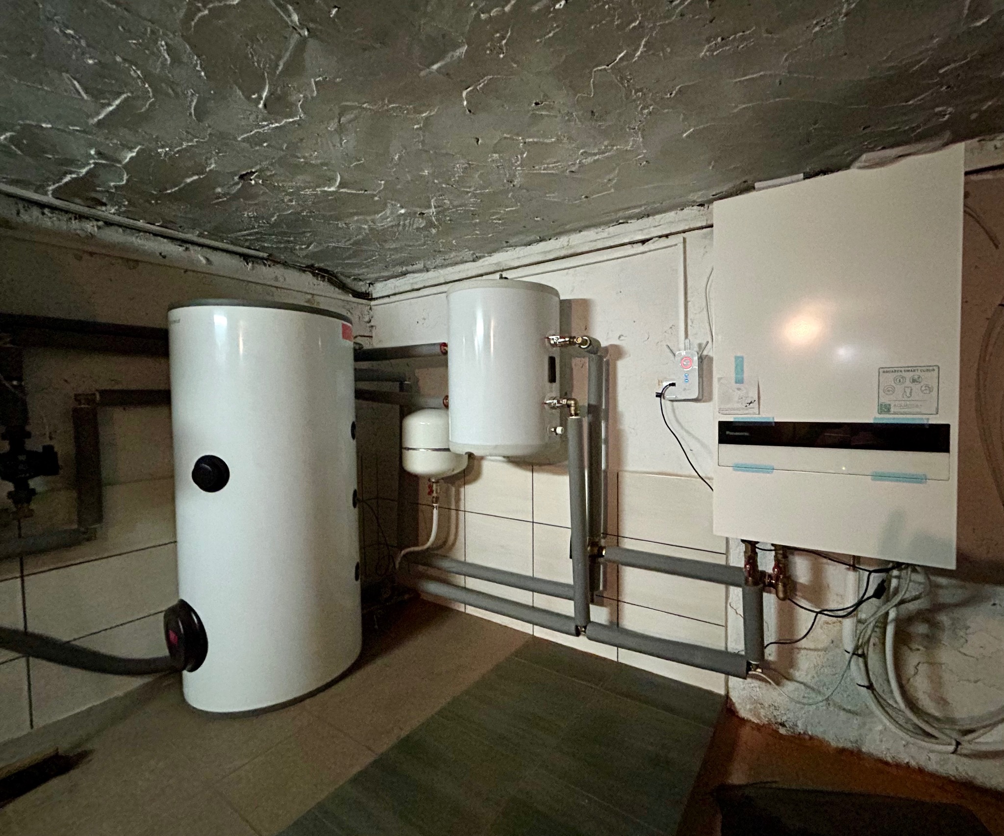 Biały bojler i piec grzewczy na tle ściany z białych płytek i szarej, chropowatej ściany. Widoczne rury instalacji grzewczej i elementy podłączenia elektrycznego.