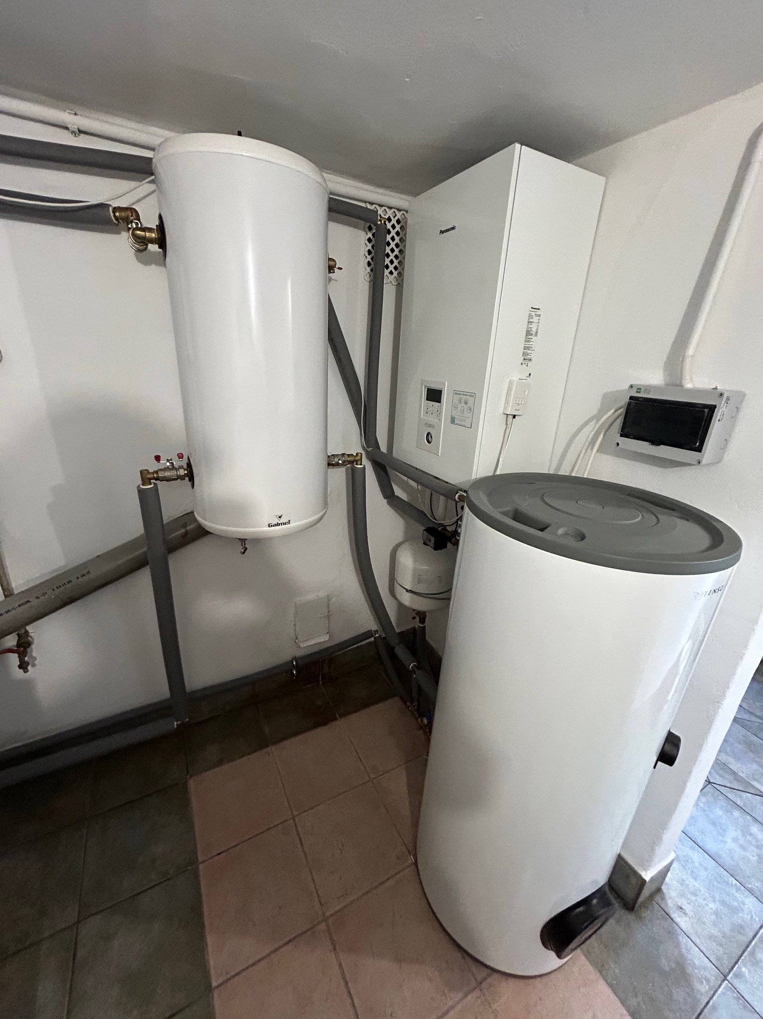 Biała pompa ciepła Galmet i zasobnik Rotenso w kotłowni. Szare rury, płytki podłogowe w odcieniach brązu. Widoczny panel sterowania Panasonic.