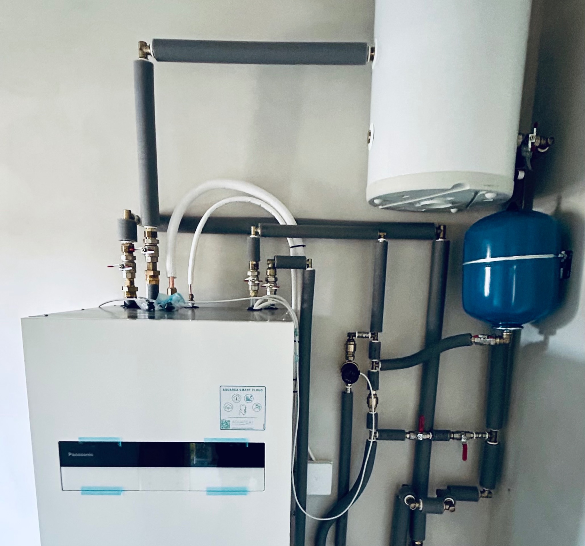 Biała pompa ciepła Panasonic Aquarea Smart Cloud z szarymi rurami izolowanymi, niebieskim zbiornikiem wyrównawczym i białym zasobnikiem ciepłej wody.