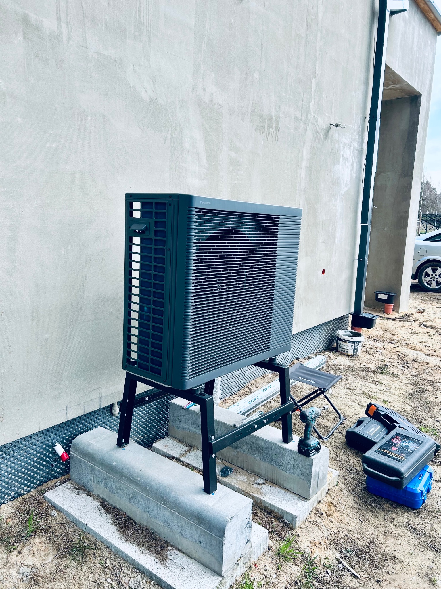Zewnętrzna jednostka pompy ciepła Panasonic Aquarea na betonowych fundamentach, z narzędziami obok, przed budynkiem z szarą elewacją. Widoczne elementy instalacji.