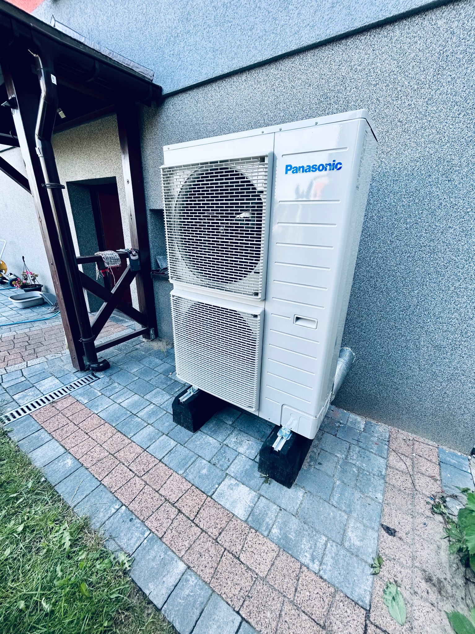 Biała, dwuczęściowa pompa ciepła Panasonic zamontowana na zewnątrz budynku na kostce brukowej, z widoczną elewacją w tle i fragmentem zadaszenia.