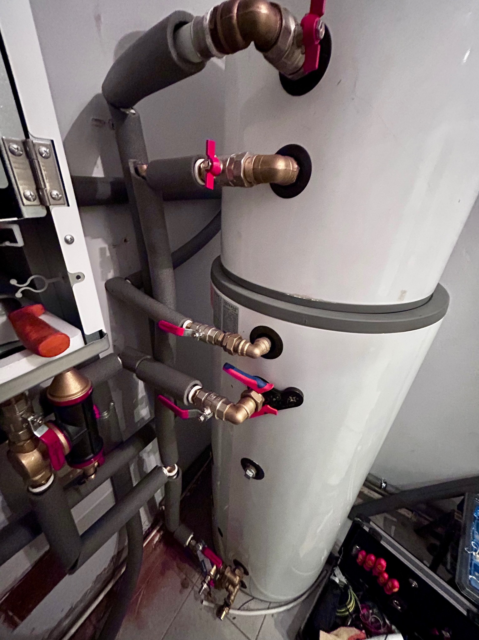 Biały, dwuczęściowy zasobnik ciepłej wody użytkowej z rurami w izolacji szarej, z zaworami z czerwonymi motylkami, zamontowany w pomieszczeniu technicznym.