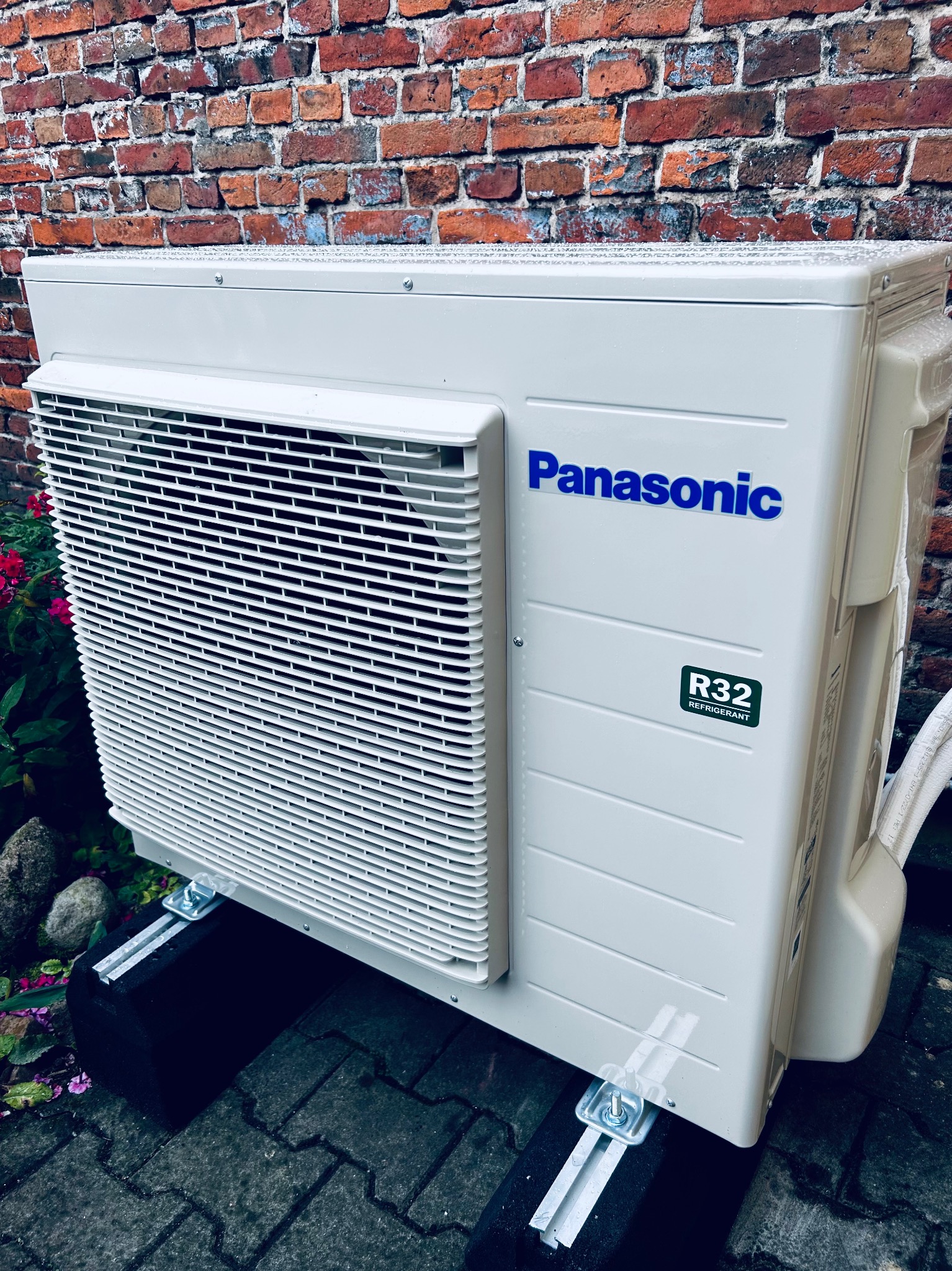 Biała jednostka zewnętrzna klimatyzacji Panasonic R32 na tle ceglanej ściany i zieleni. Widoczne detale montażu na czarnych podkładkach i metalowych wspornikach.