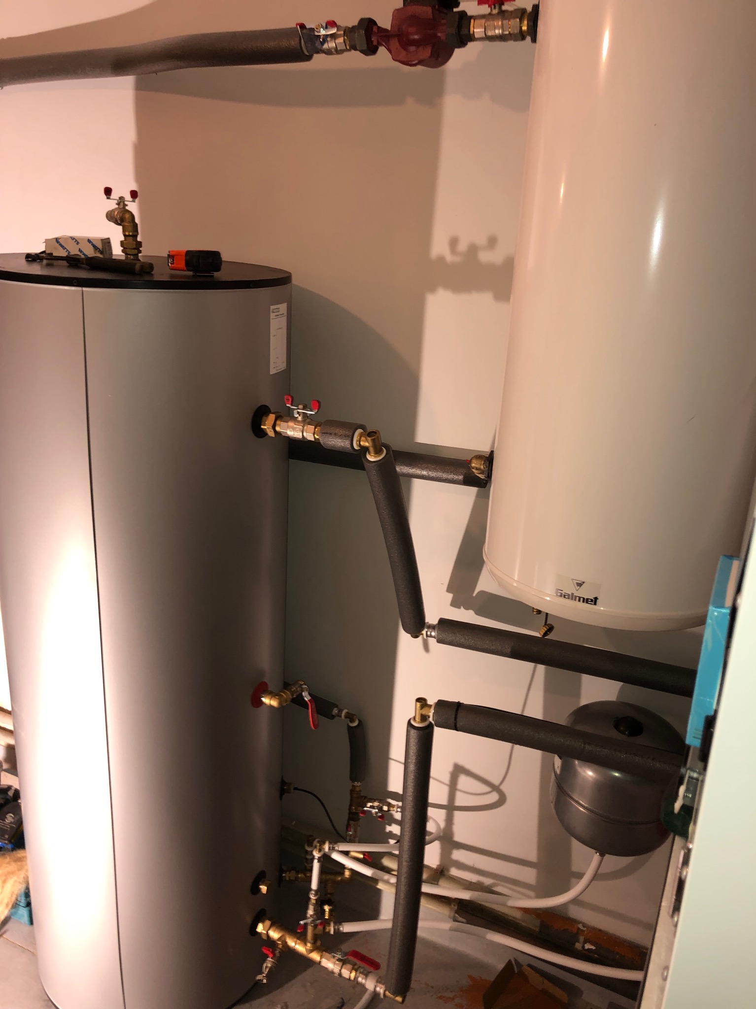 Dwa zbiorniki, w tym jeden marki Galmet, połączone rurami z izolacją termiczną, w pomieszczeniu technicznym. Widoczne zawory i elementy instalacji grzewczej.