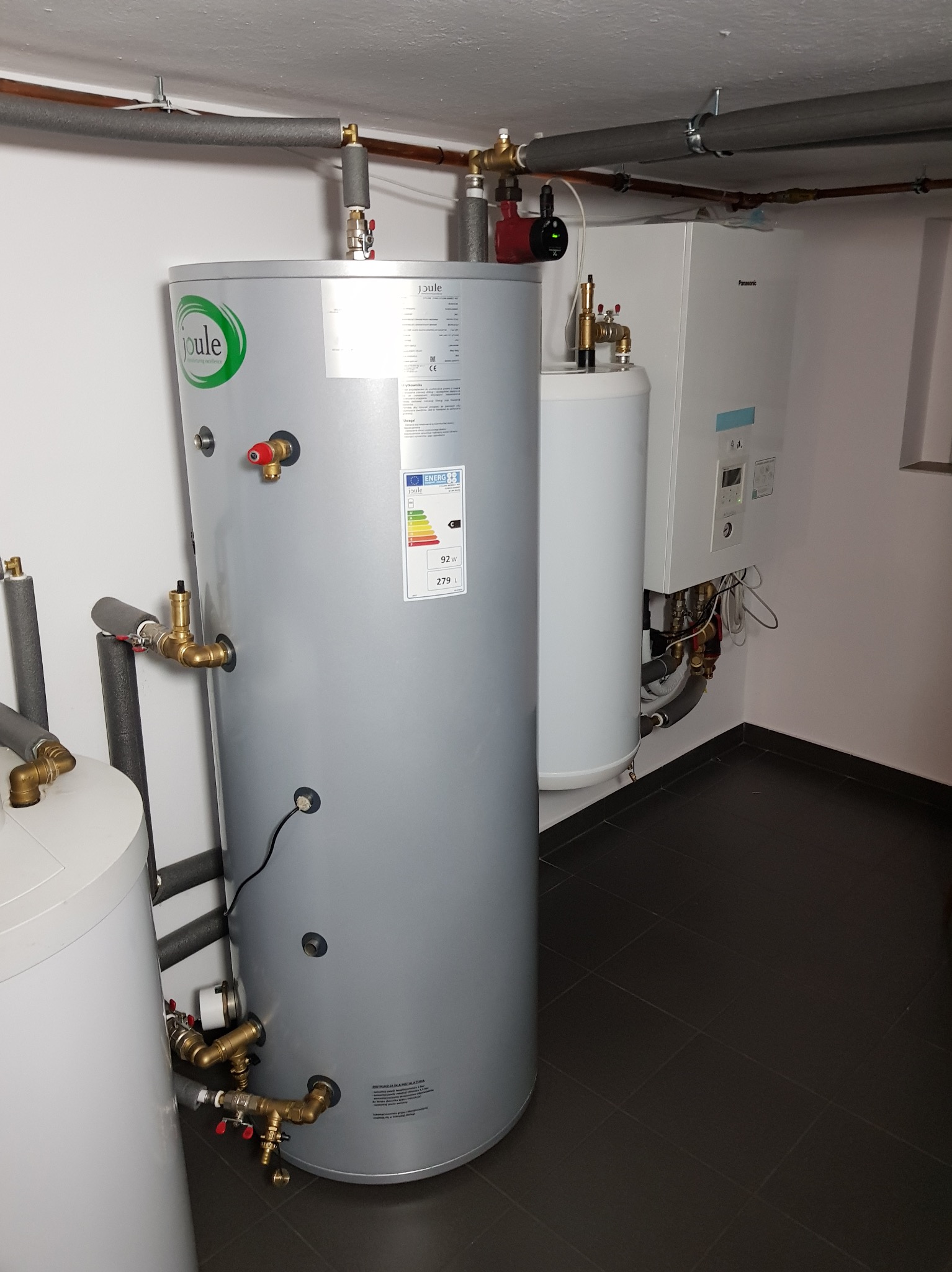 Szary, cylindryczny zbiornik Joule z etykietą energetyczną, obok biały bojler Panasonic, rury i czarna podłoga z płytek. Instalacja w pomieszczeniu.