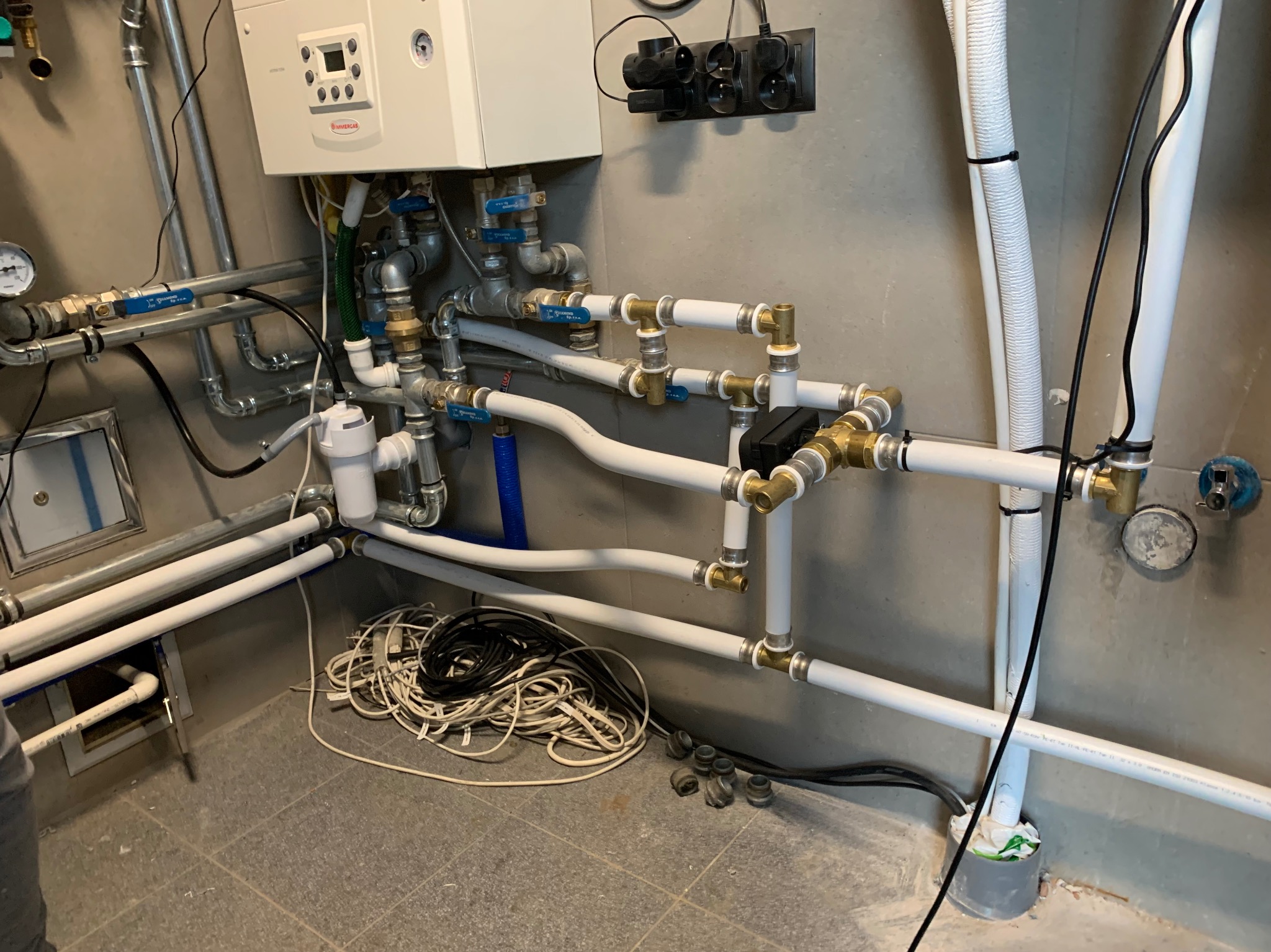 Instalacja grzewcza z kotłem gazowym, rurami z zaworami i przewodami elektrycznymi na tle szarej ściany. Widoczne elementy instalacji w pomieszczeniu technicznym.