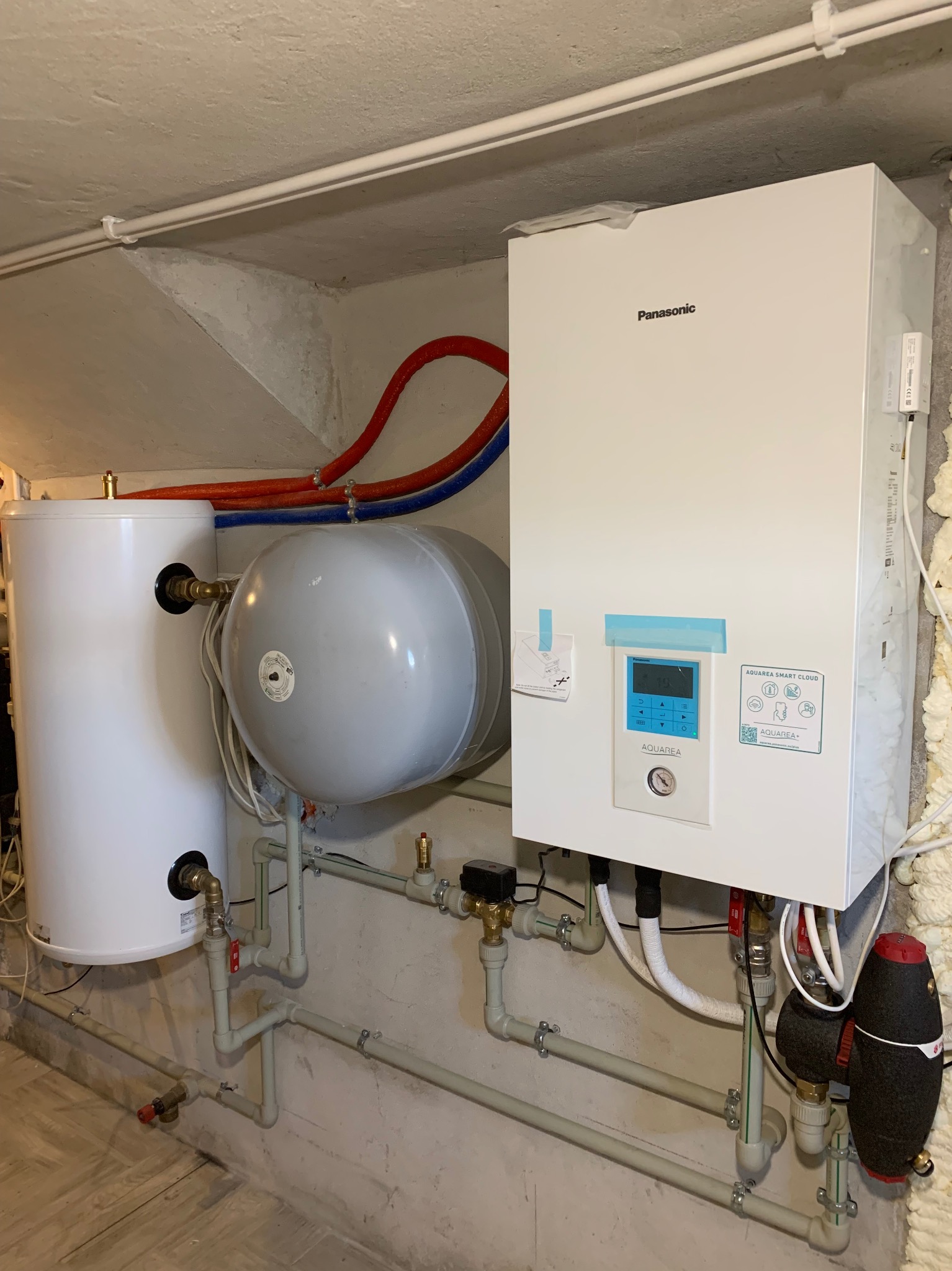 Zestaw instalacji grzewczej: biały zbiornik, szary zbiornik ciśnieniowy i pompa ciepła Panasonic Aquarea Smart Cloud z rurami w piwnicy.
