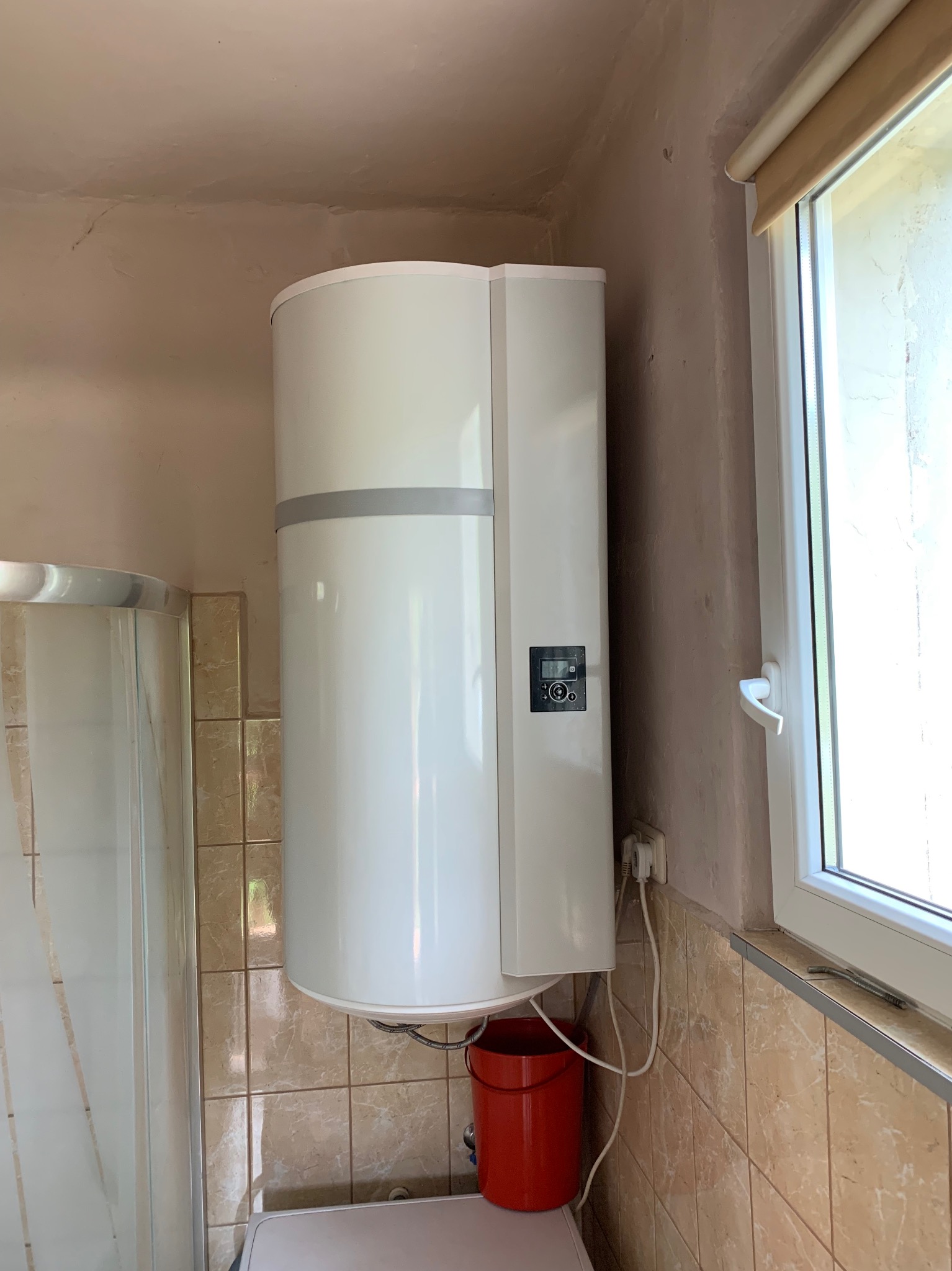 Biała pompa ciepła w łazience z beżowymi płytkami, widoczny fragment kabiny prysznicowej, okno i czerwone wiadro. Ujęcie z dołu, widoczne niedoskonałości ścian.