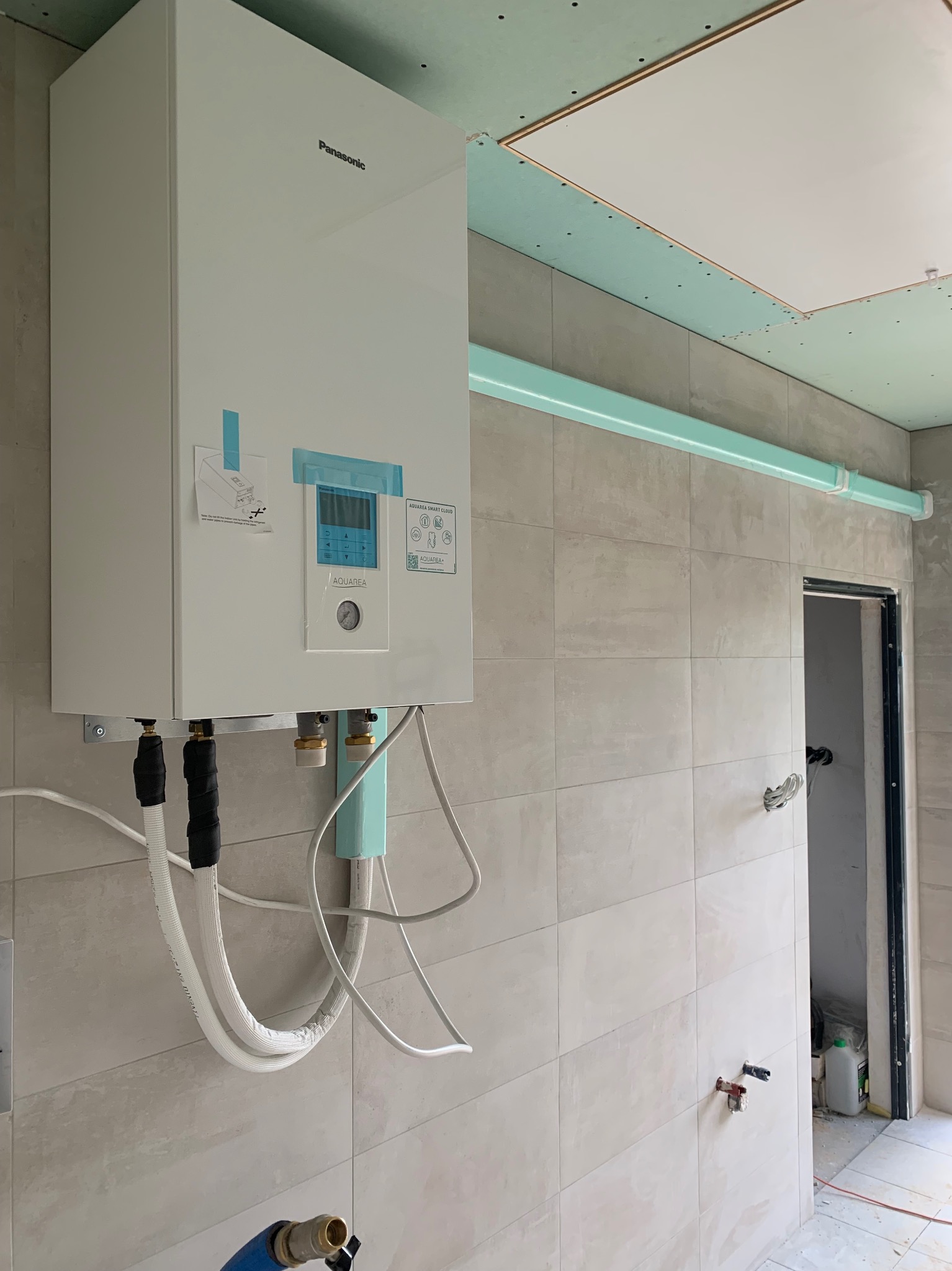 Biała pompa ciepła Panasonic Aquarea zamontowana na ścianie z szarych płytek, widoczne rury i instalacja, sufit z płyt kartonowo-gipsowych w trakcie montażu.