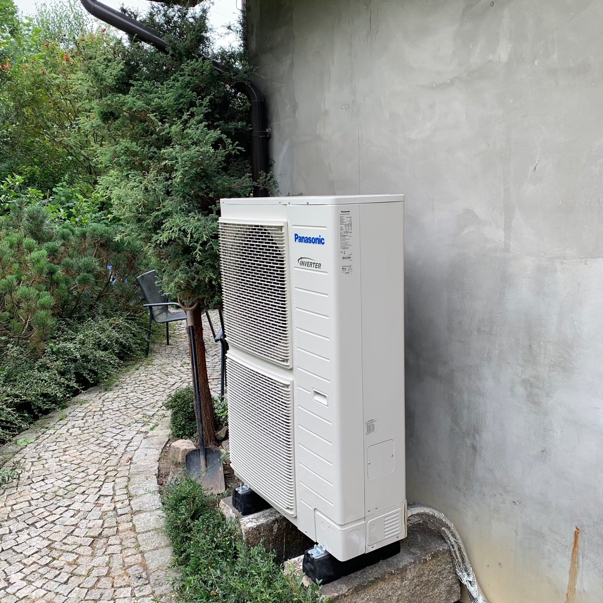 Zewnętrzna jednostka klimatyzacji Panasonic Inverter, zamontowana na betonowym podeście przy budynku z szarą elewacją. Obok ścieżka z kostki i zieleń.