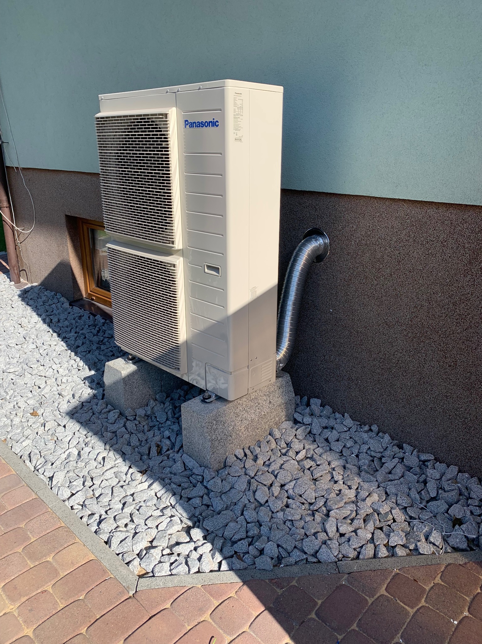 Zewnętrzna jednostka klimatyzacji Panasonic zamontowana na betonowych podstawach, z widocznym przewodem odprowadzającym skropliny. Otoczenie wyłożone ozdobnym kamieniem.